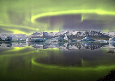 A vivid green overheaded aurrora pictured in Iceland's Vatnajokull National Park reflected almost symetrically in Jokulsrlon Glacier lagoon. A complete lack of wind and currrent combin in this sheltred lagoon scene to crete an arresting mirror effect giving the image a sensation of utter stillness. Despite theis there is motion on a suprising scale, as the loops and arcs of the aurora are shaped by the shifting forces of the Earth's magnetic field.