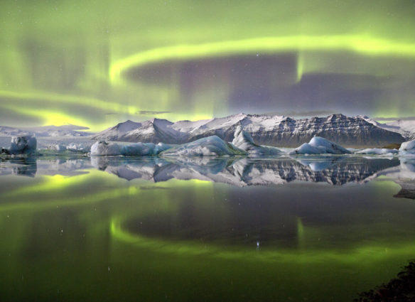 A vivid green overheaded aurrora pictured in Iceland's Vatnajokull National Park reflected almost symetrically in Jokulsrlon Glacier lagoon. A complete lack of wind and currrent combin in this sheltred lagoon scene to crete an arresting mirror effect giving the image a sensation of utter stillness. Despite theis there is motion on a suprising scale, as the loops and arcs of the aurora are shaped by the shifting forces of the Earth's magnetic field.